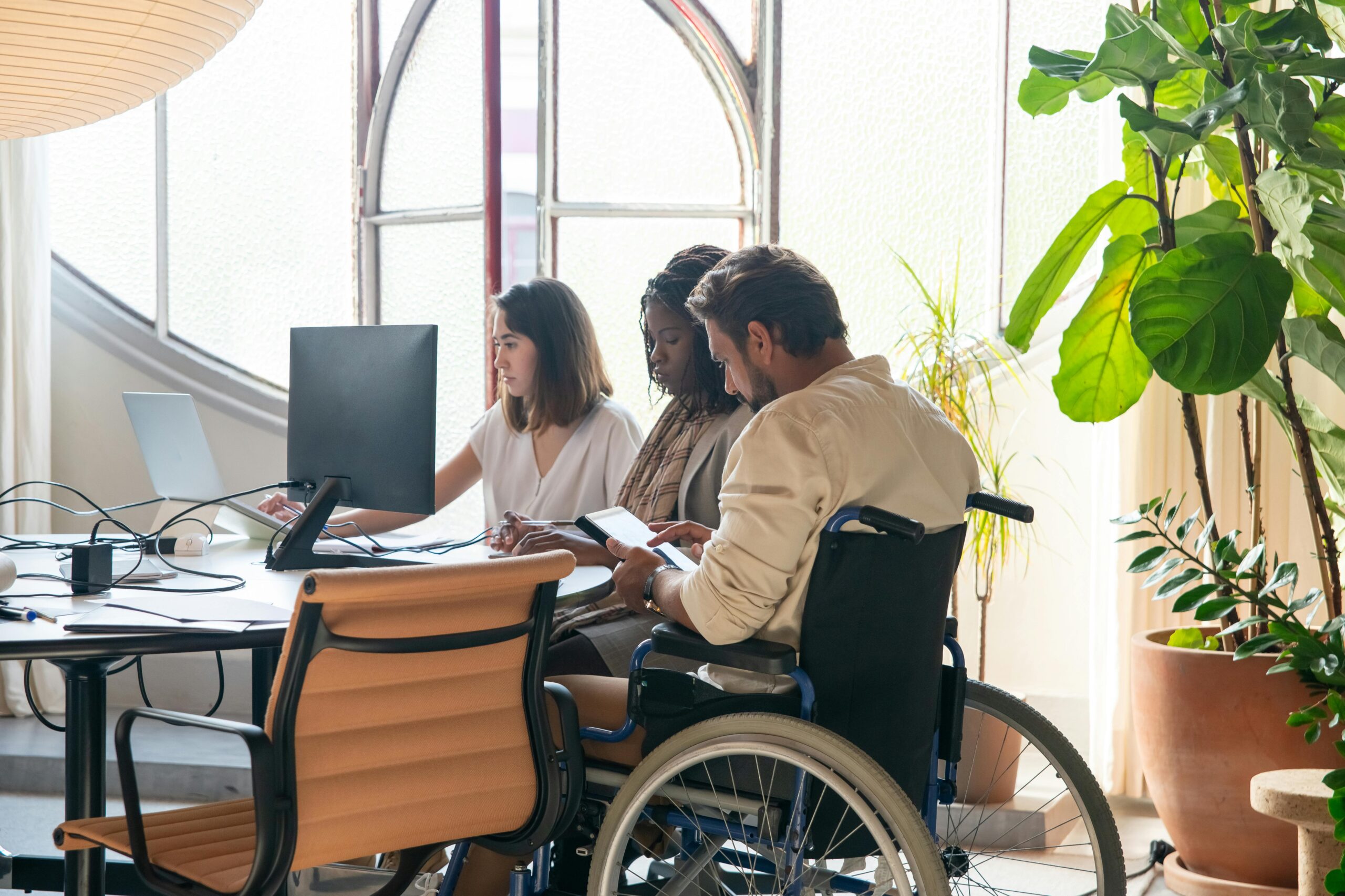 Three colleagues working together at a shared desk in a bright, plant-filled office, including a man using a wheelchair who is reviewing content on a tablet. Photo by Kampus Production from Pexels.