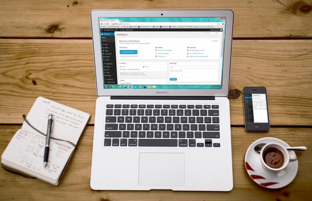 An open silver MacBook Air displays a WordPress welcome dashboard on a wooden workspace, flanked by a pen on a handwritten notebook, a smartphone showing a mobile screen, and an espresso cup on a saucer.
