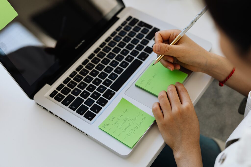 Over-the-shoulder view of a person holding a pen and writing on a green sticky note placed on a laptop keyboard base next to another handwritten list.