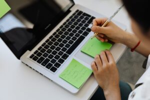 Over-the-shoulder view of a person holding a pen and writing on a green sticky note placed on a laptop keyboard base next to another handwritten list.