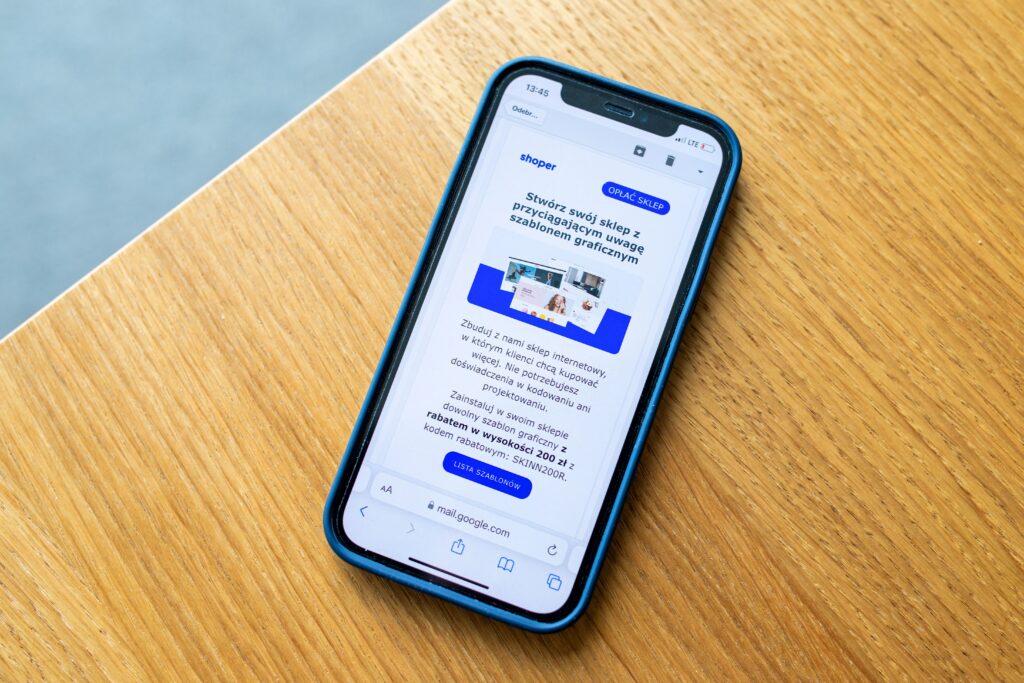 Top-down view of a smartphone in a blue protective case sitting on a light oak wooden table. The phone screen displays an open email in a mobile browser featuring the blue and white Shoper logo, headline text in Polish, and a promotional offer with a blue button at the bottom.
