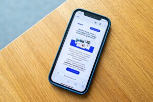 Top-down view of a smartphone in a blue protective case sitting on a light oak wooden table. The phone screen displays an open email in a mobile browser featuring the blue and white Shoper logo, headline text in Polish, and a promotional offer with a blue button at the bottom.