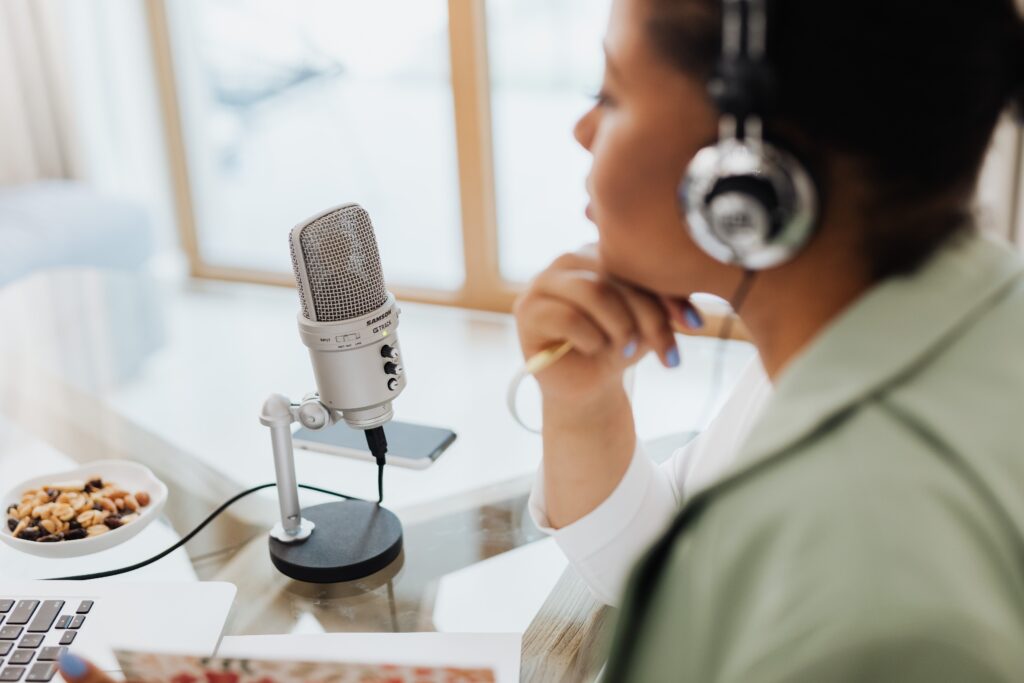 A silver Samson G-Track studio condenser microphone mounted on a small desktop stand sits in sharp focus on a glass-top table. A laptop keyboard, a small dish of trail mix or nuts, and a smartphone are nearby on the desk.
